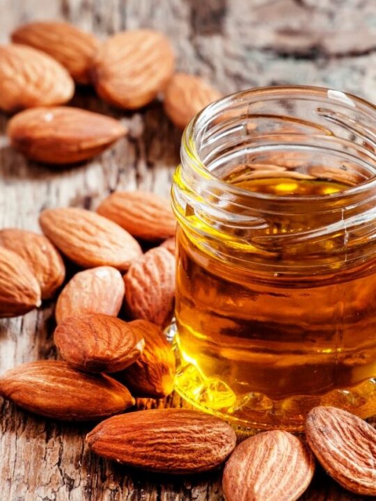 Clear glass jar of almond extract surrounded by almonds on a wooden table.