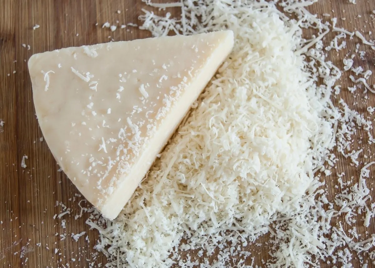 Large wedge of parmesan cheese on wooden cutting board next to grated cheese.