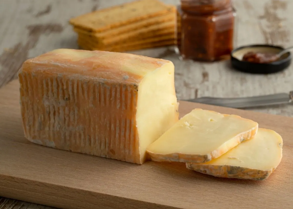Large block of taleggio cheese next to slices on a wooden cutting board with crackers and jam in background.