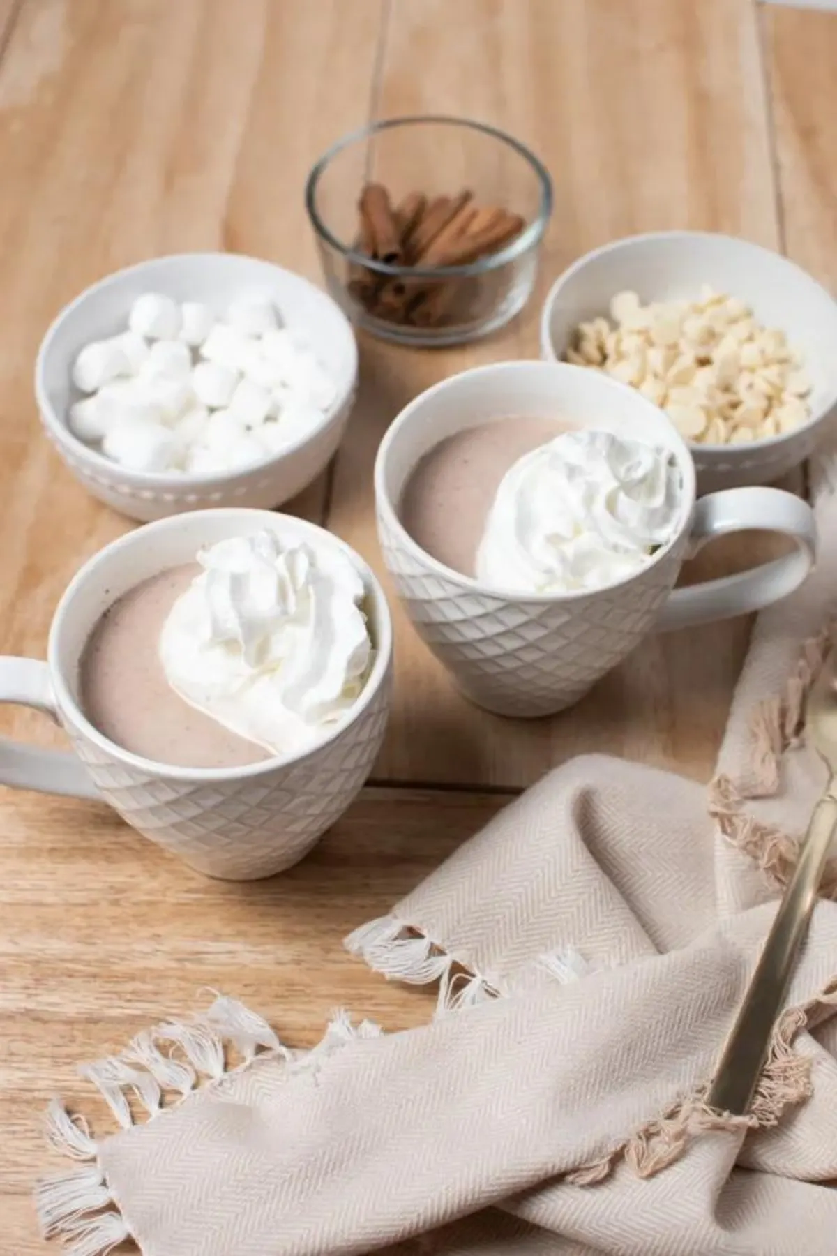 Mugs filled with white hot chocolate next to bowls of chocolate, marshmallow and cinnamon.