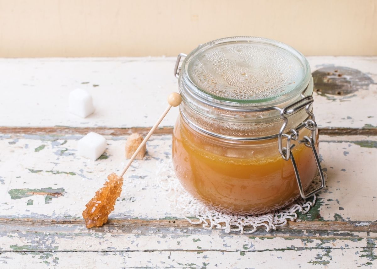 Brown rice syrup in a glass jar next to a stick of rock sugar on rustic wood table.