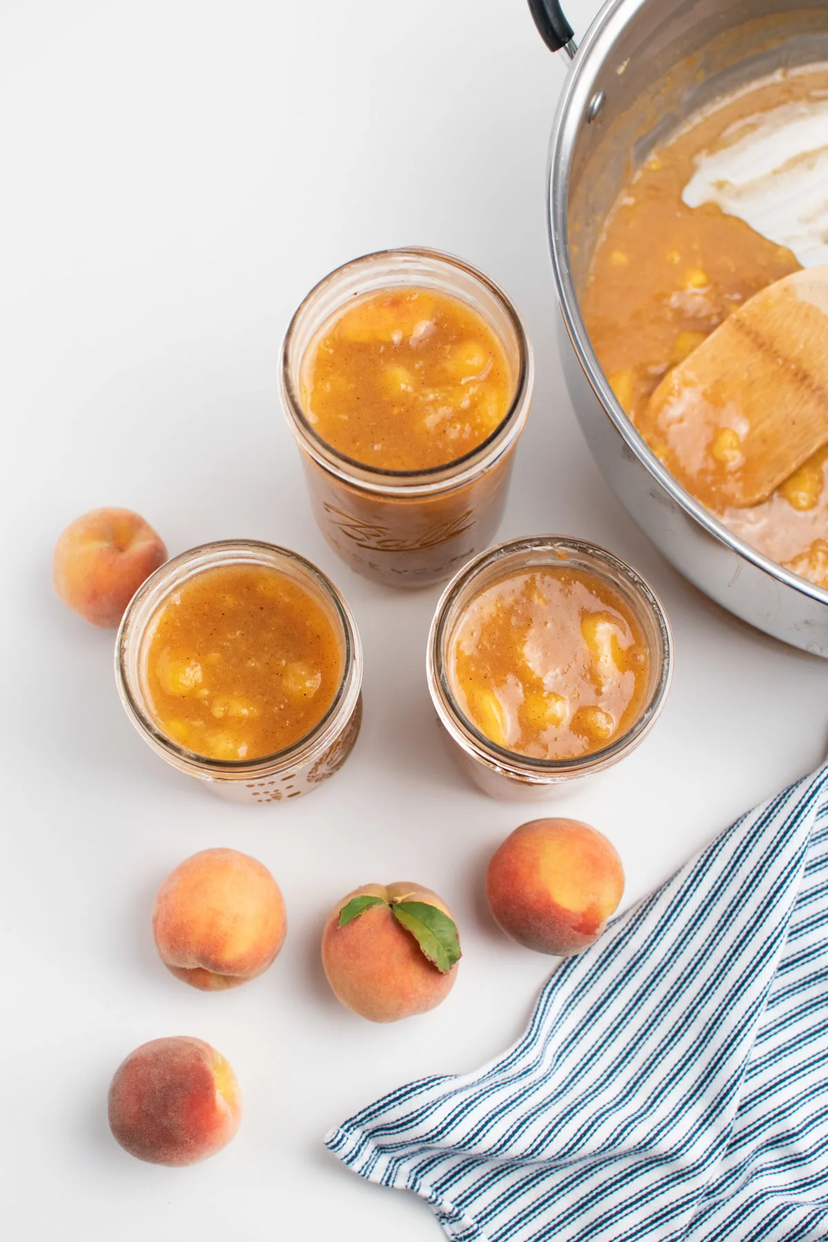 Three jars of peach pie filling surrounded by fresh peaches and striped towel.