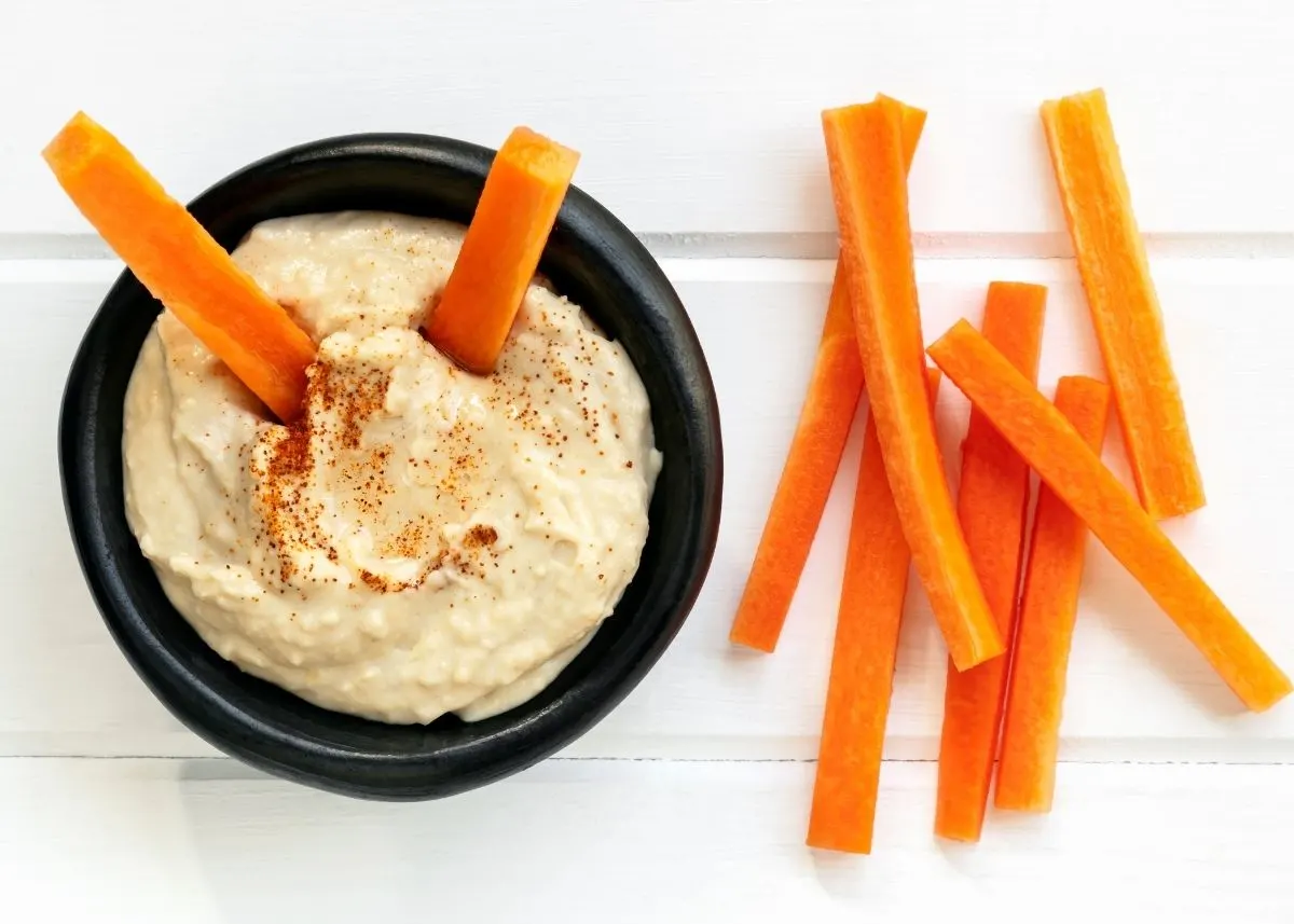 Two carrots stick out of a bowl of hummus next to carrots on a table.