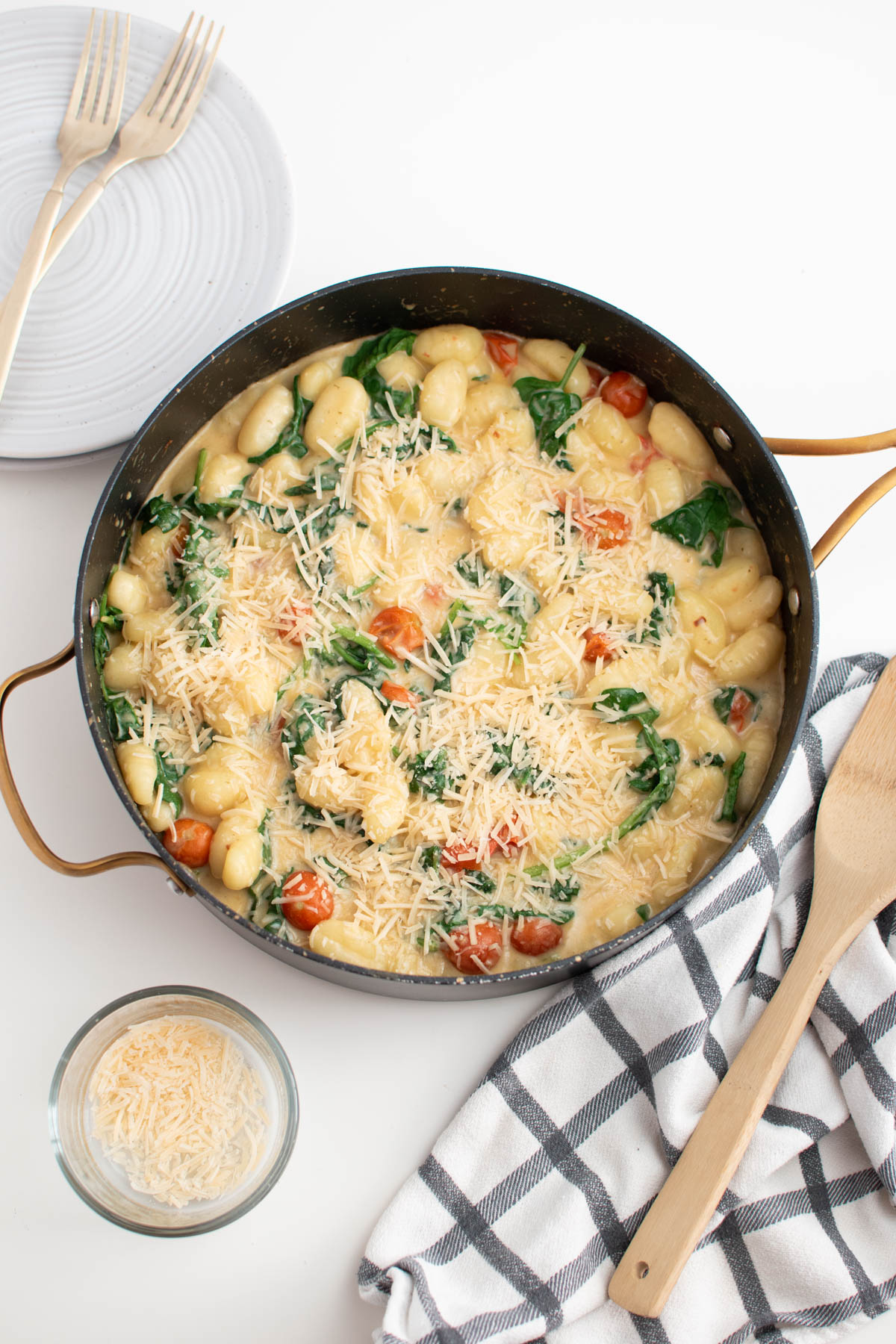 Large skillet of creamy gnocchi with tomatoes surrounding by wooden spoon and plates on white table.