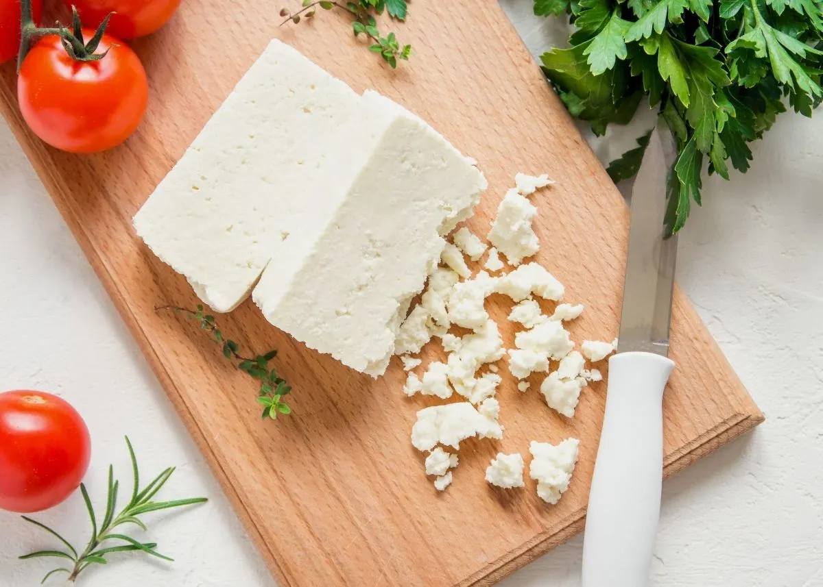 Two blocks of feta cheese on a wooden cutting board next to crumbles.