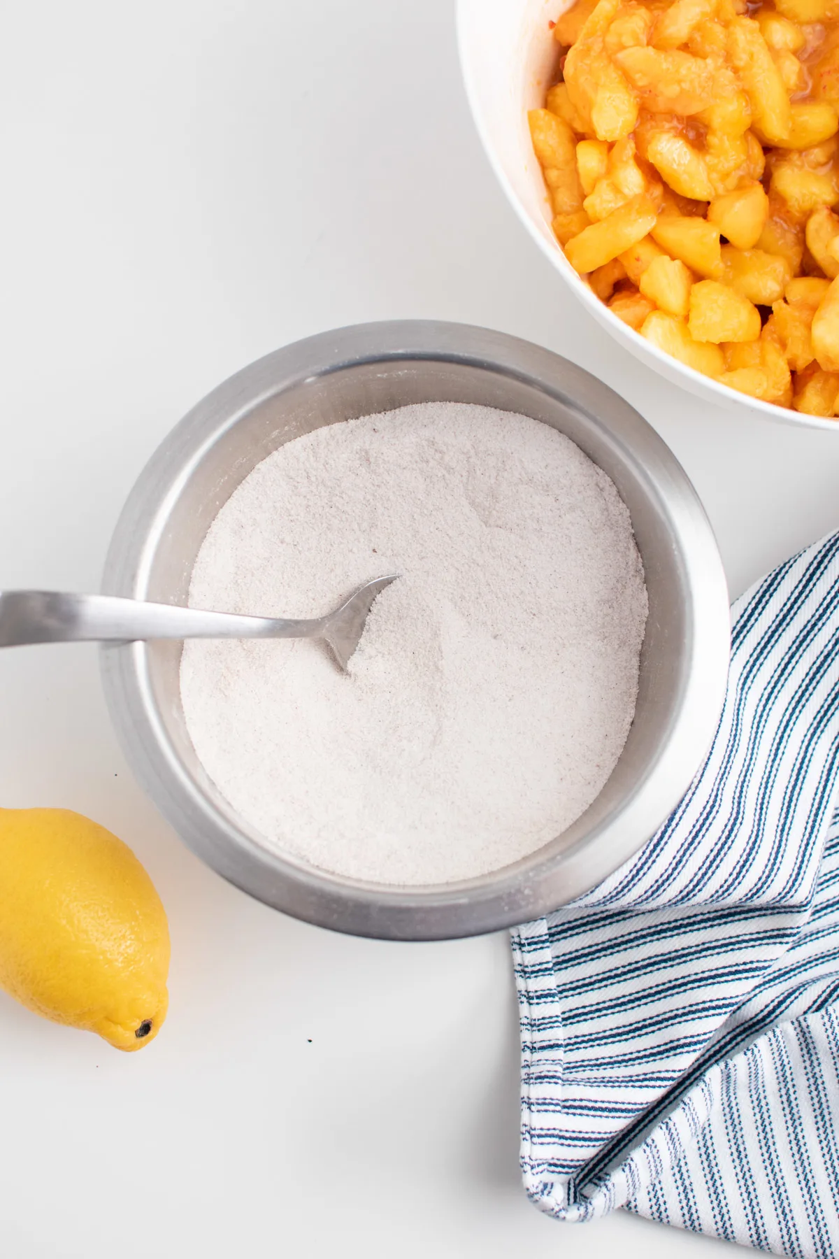 Flour and sugar mixture in small bowl with spoon surrounded by towel, lemon, and sliced peaches.