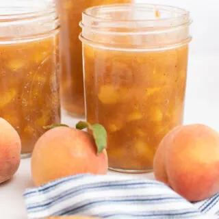 Three jars of freezer peach pie filling surrounded by fresh peaches and striped towel.