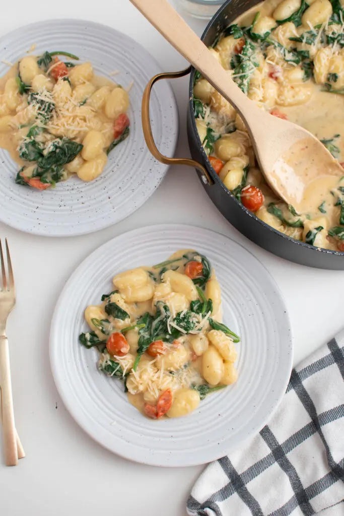 Two plates and large skillet with creamy gnocchi and tomatoes all on white table.