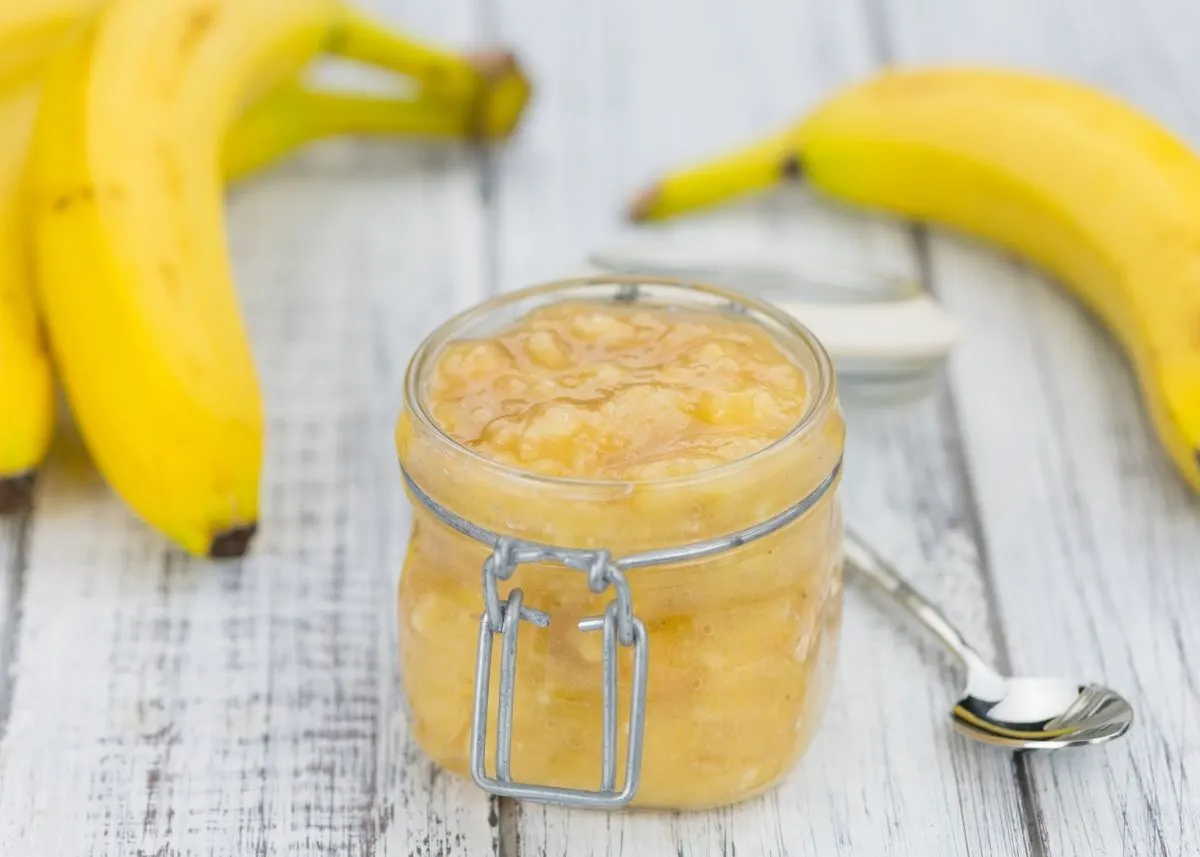 Jar of mashed, ripened bananas made into syrup next to three unpeeled bananas.