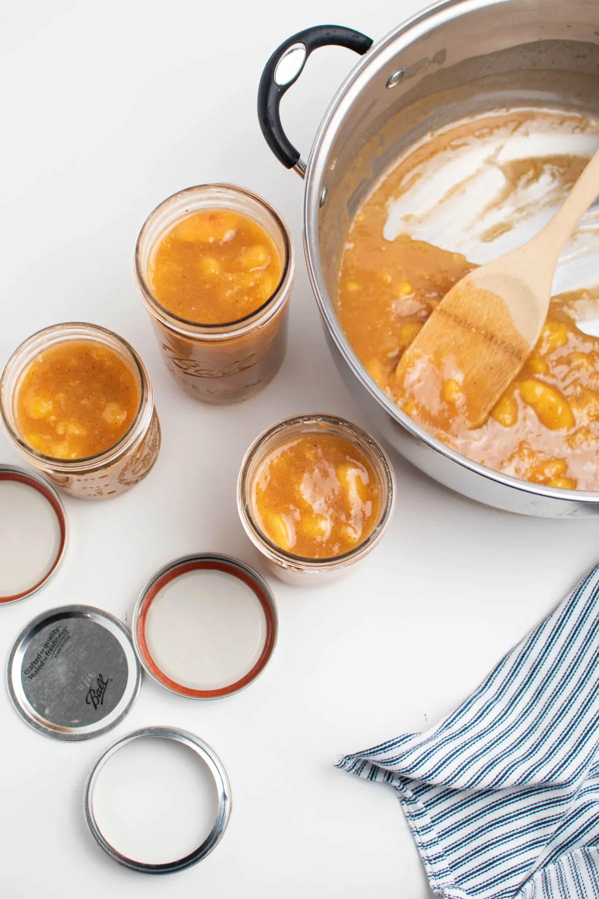 Three jars full of peach pie filling surrounded by jar lids, towel, and pot of pie filling.