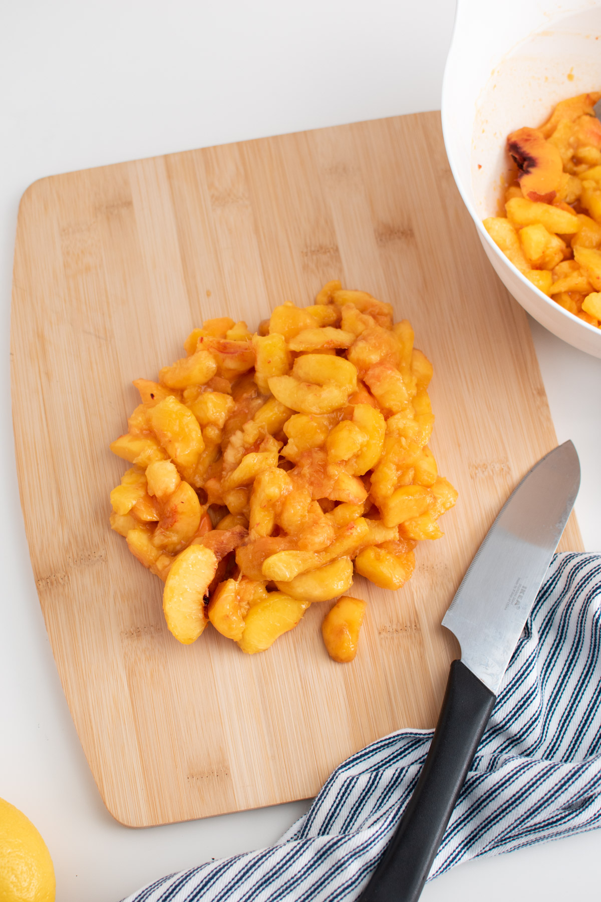 Large pile of sliced peaches on cutting board surrounded by knife, striped towel, and bowl of peaches.