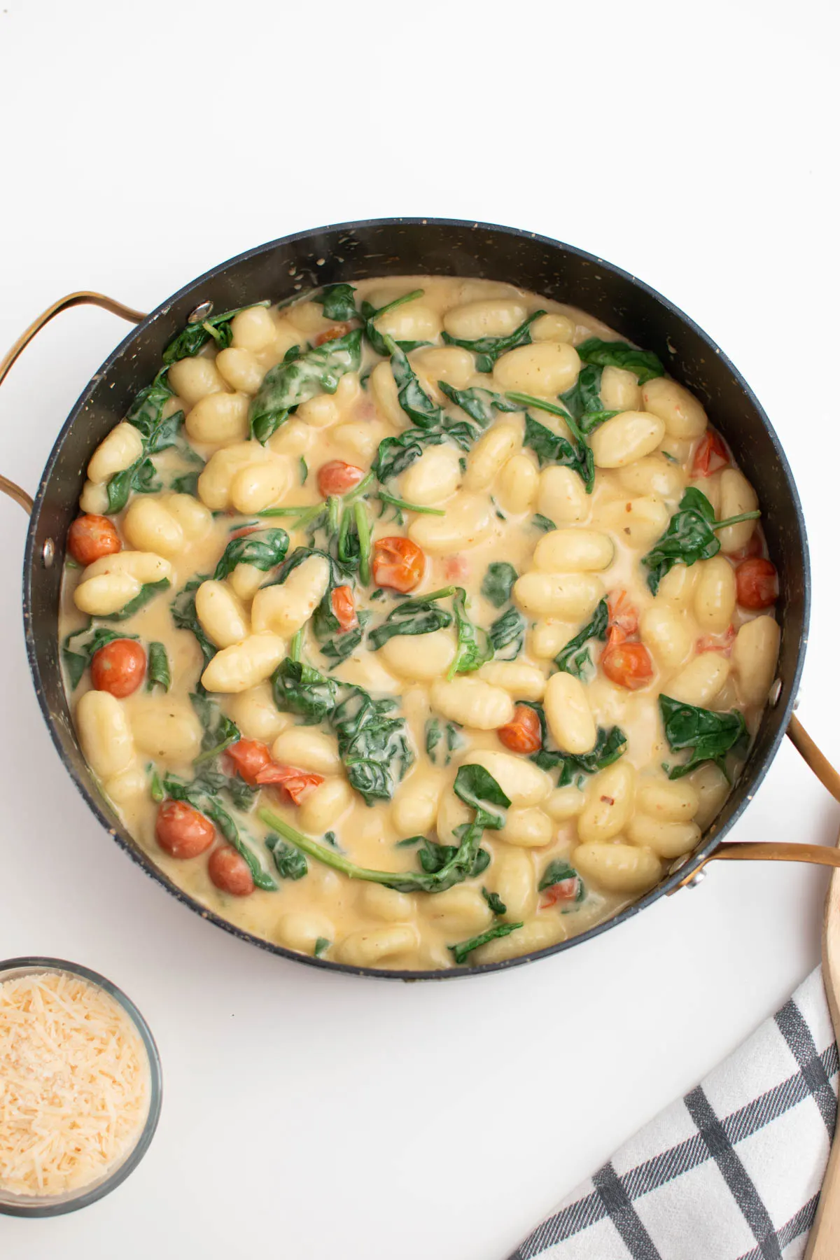 Large black skillet full of gnocchi, tomatoes, and spinach next to bowl of parmesan cheese.