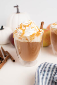 Glass mug of caramel apple cider with whipped cream in front of pumpkins and apples.
