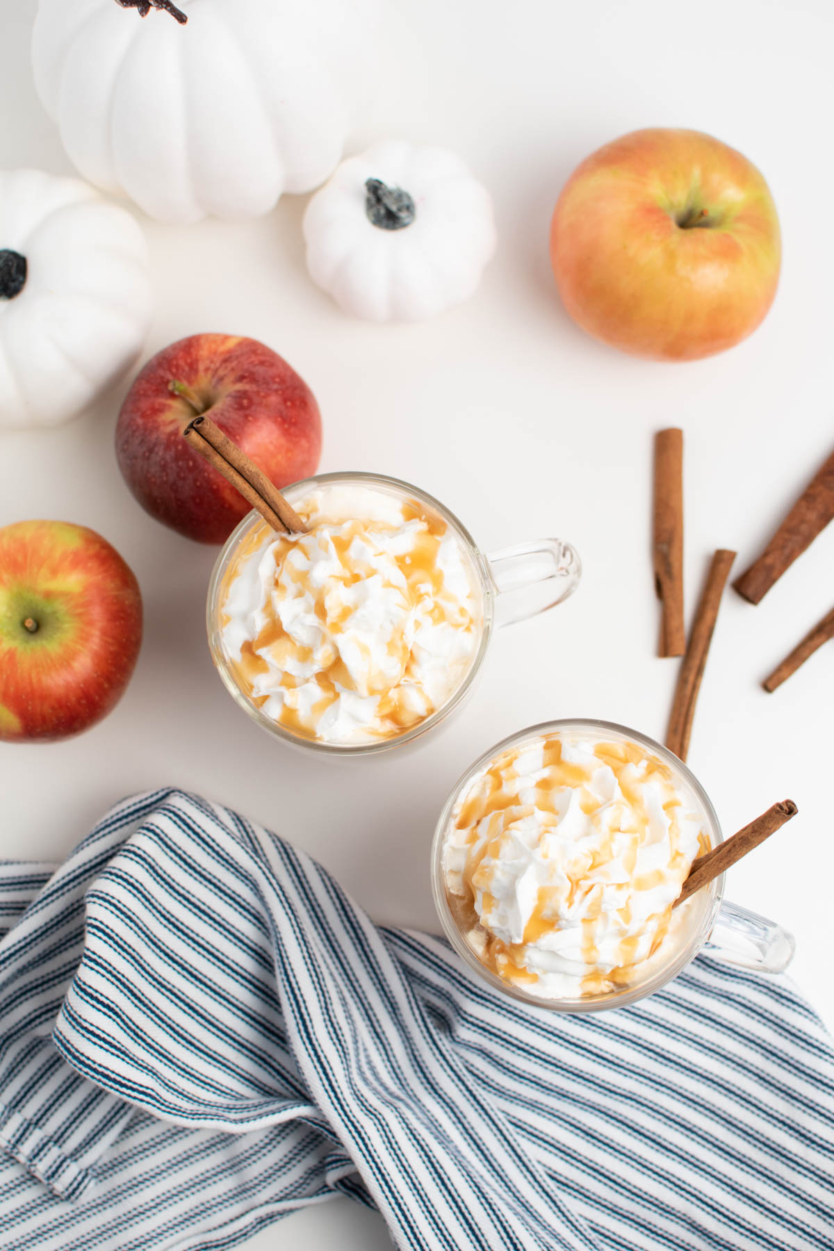 Two mugs of caramel apple cider with whipped cream on white table with fall decorations.