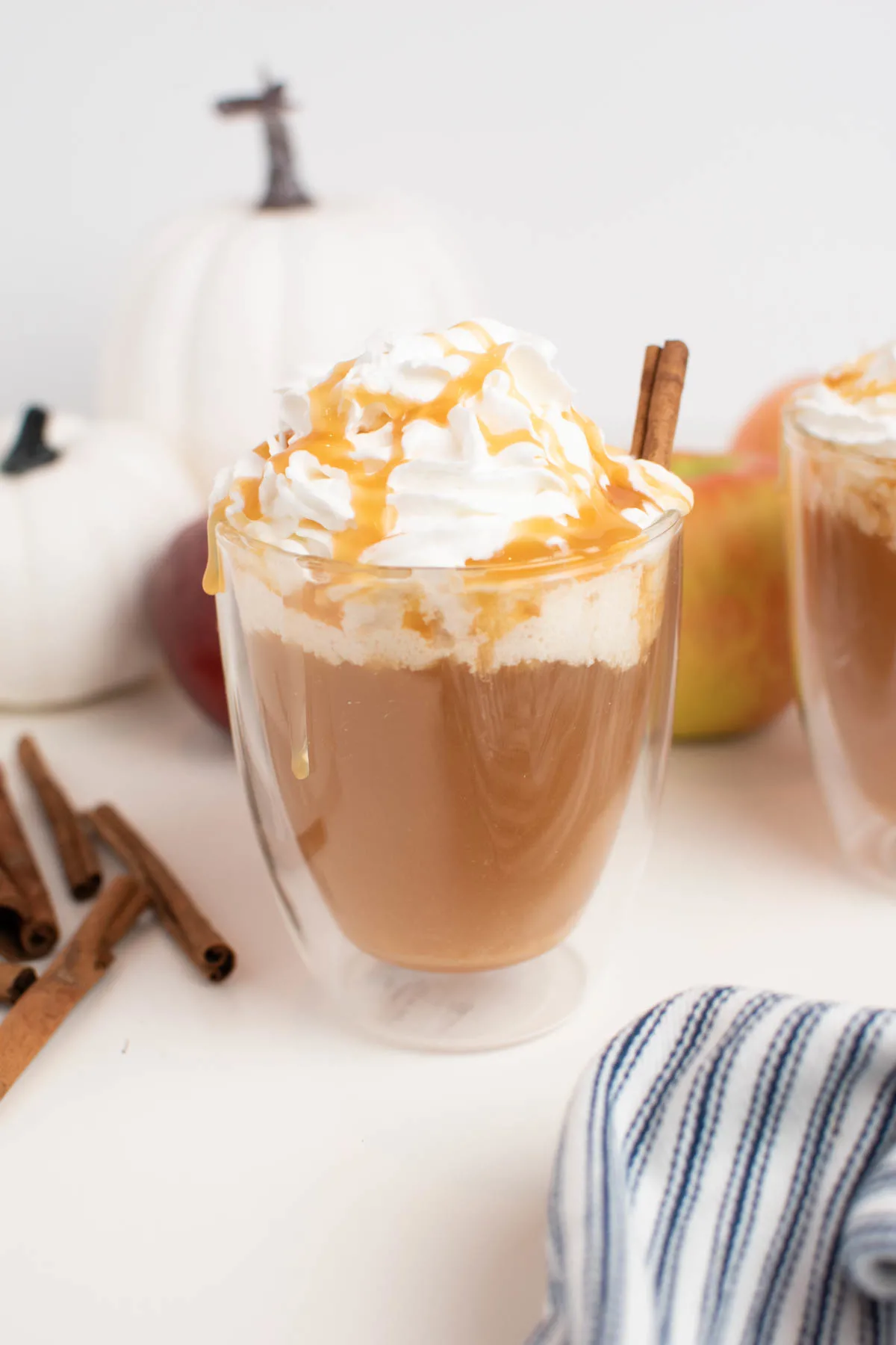 Glass mug of caramel apple cider with whipped cream in front of pumpkins and apples.