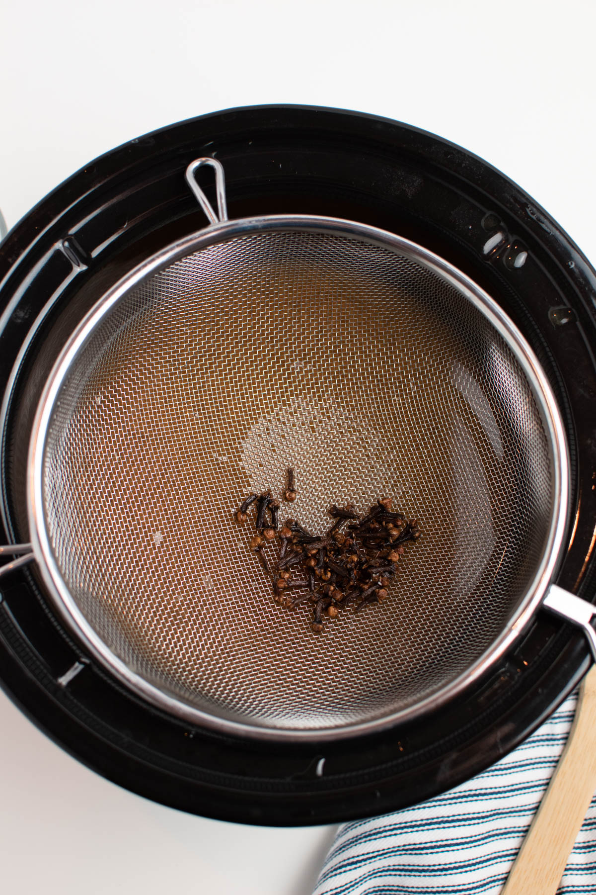 Fine mesh sieve filled with whole cloves over black slow cooker insert.