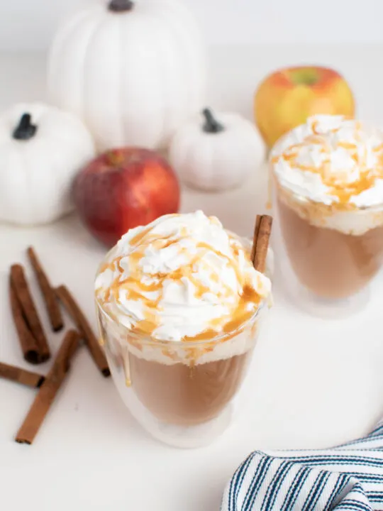 Two mugs of hot caramel apple cider on table surrounded by fall decorations.