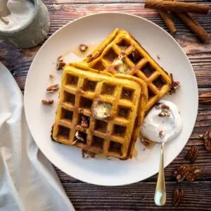Two pumpkin waffles on a white plate with melting butter on top and chopped nuts.