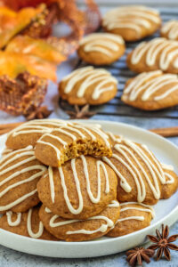 Several pumpkin spice cookies with zigzag frosting piled on white plate next to cooking rack.