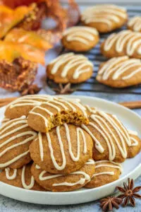 Several pumpkin spice cookies with zigzag frosting piled on white plate next to cooking rack.