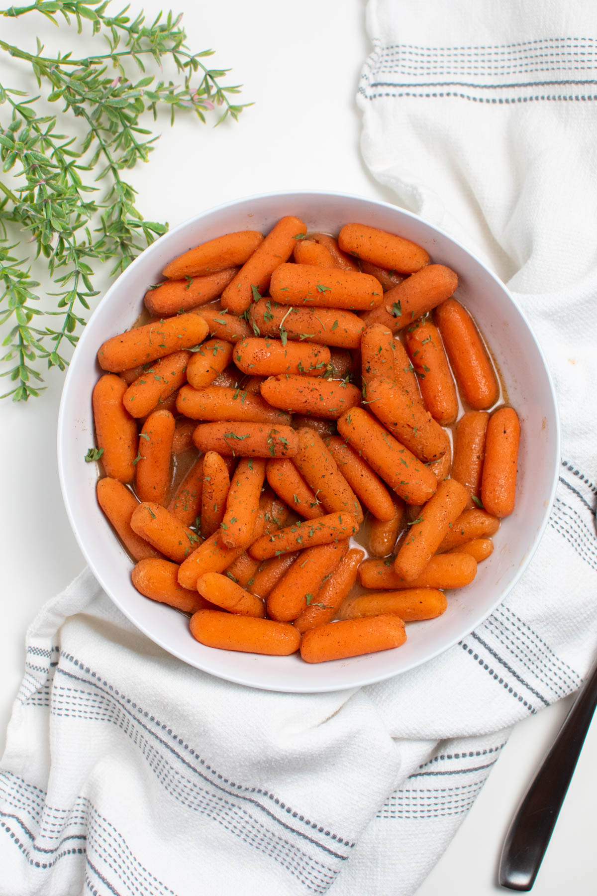 Brown sugar and honey glazed carrots in white bowl next to white towel and faux green stems.