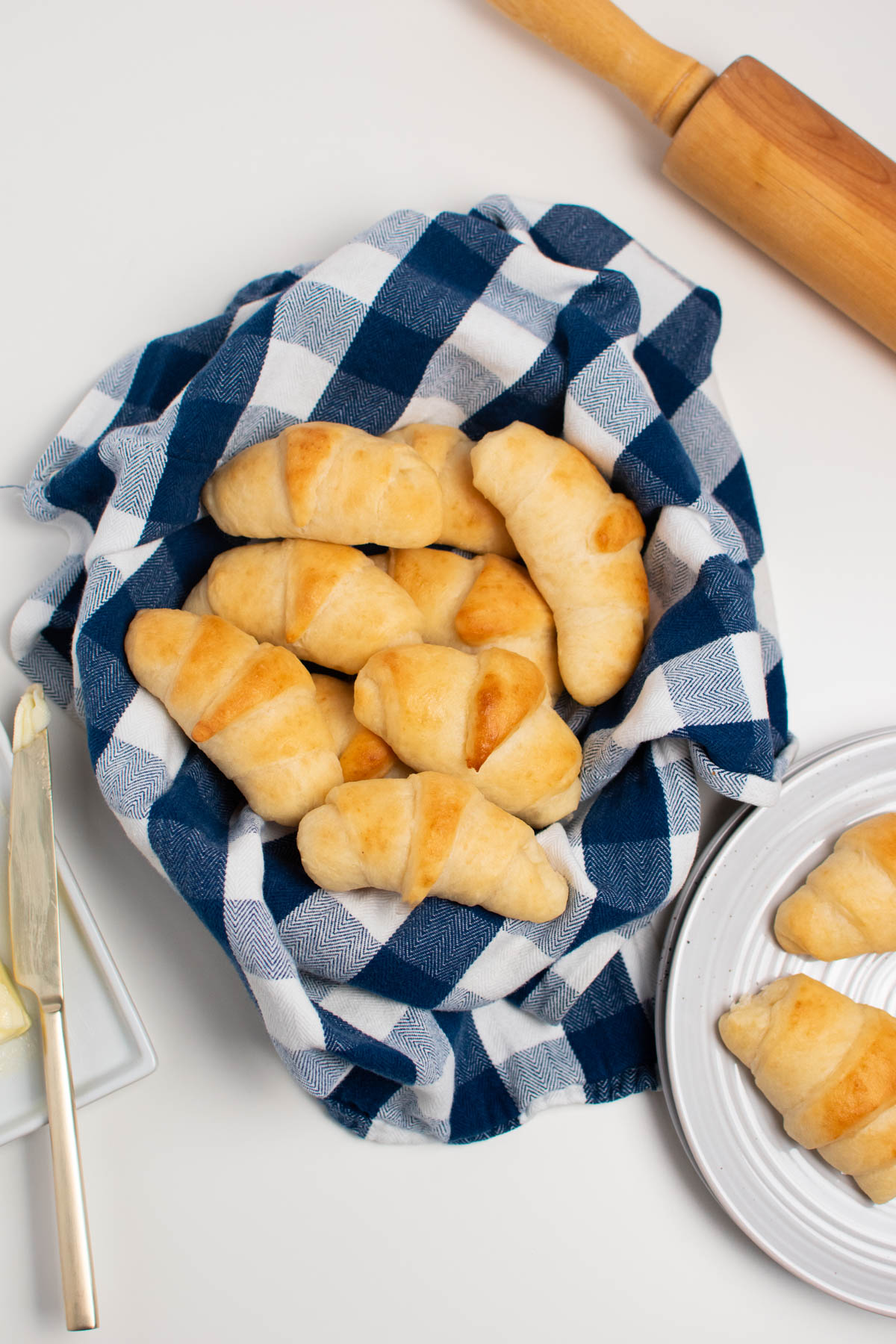 Several crescent rolls in bowl with blue checkered towel on white table.
