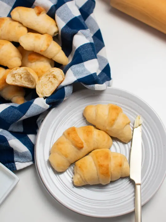 Three crescent rolls next to butter knife on white plate surrounded by bowl of rolls and rolling pin.