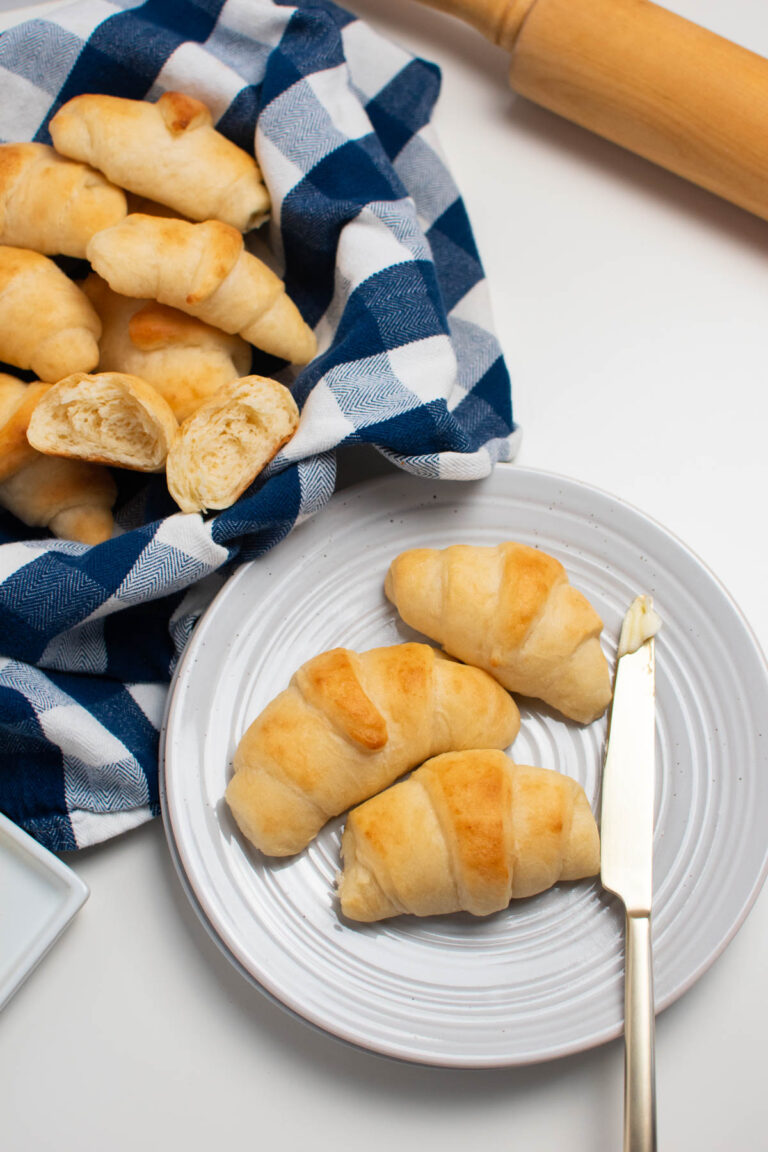 Three crescent rolls next to butter knife on white plate surrounded by bowl of rolls and rolling pin.