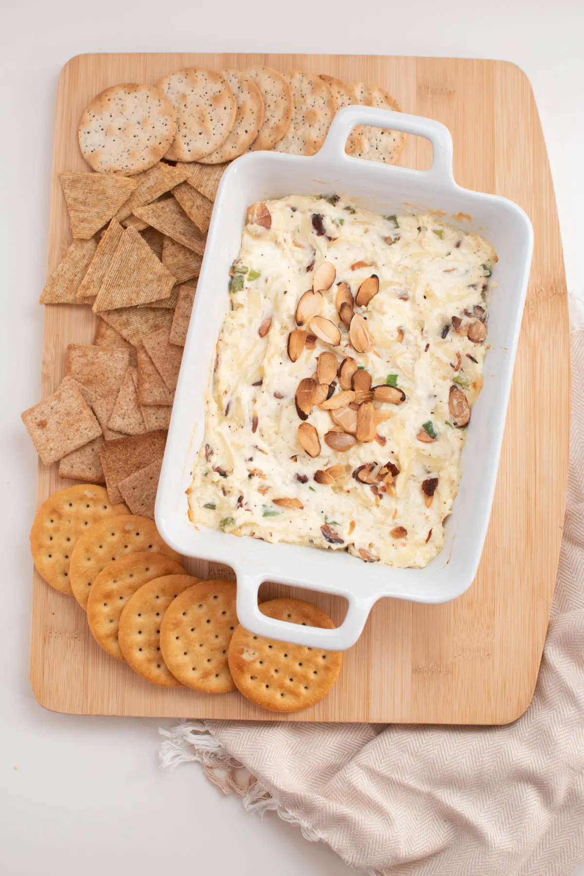 Hot almond Swiss dip in white baking dish and various crackers on wood cutting board.