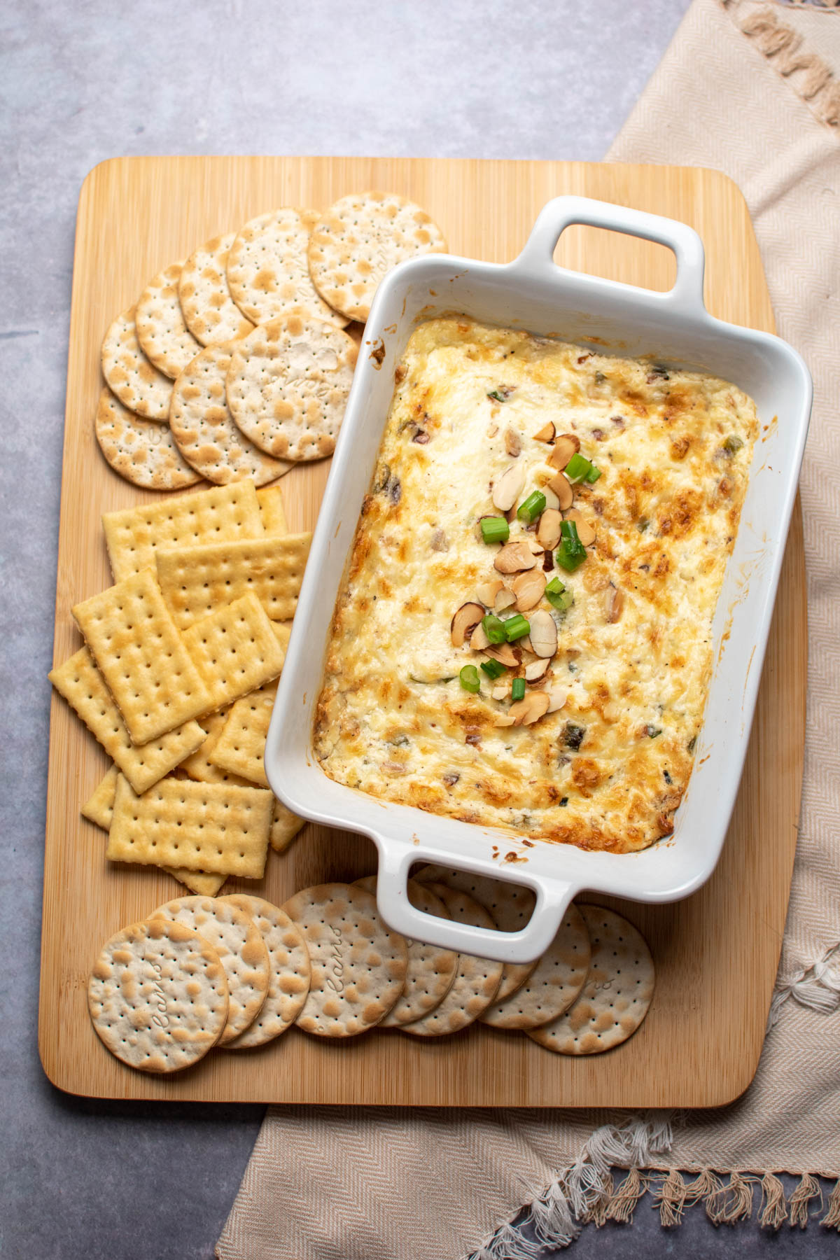 Small white baking dish with hot Swiss cheese dip and assorted crackers on wood cutting board.