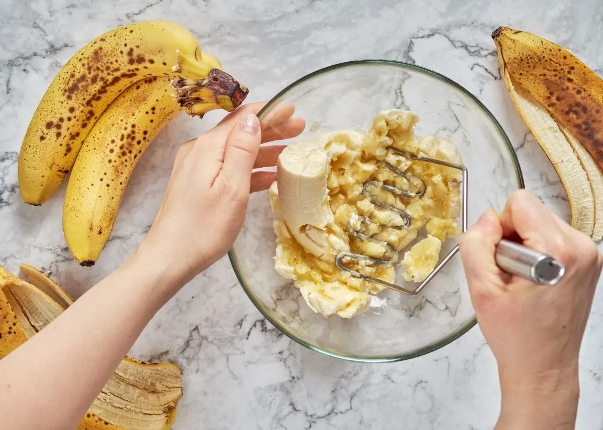 Woman mashes several bananas in large glass mixing bowl next to peels.