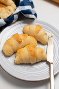Three crescent rolls and butter knife on white plate on white table.