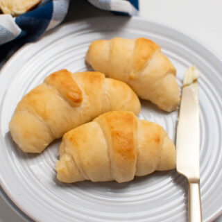 Three crescent rolls and butter knife on white plate on white table.