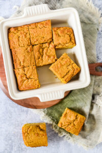 Pumpkin cornbread in small white baking dish cut into squares on wooden cutting board.