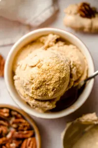 Pumpkin ice cream in a cream colored bowl with spoon next to bowl of pecans.