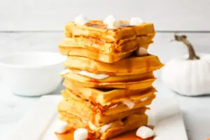 Large stack of pumpkin waffles with syrup spilling down and marshmallows sprinkled on top.