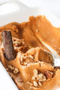 Spoon lifts pumpkin spice nice cream with chopped nuts from a white casserole dish.