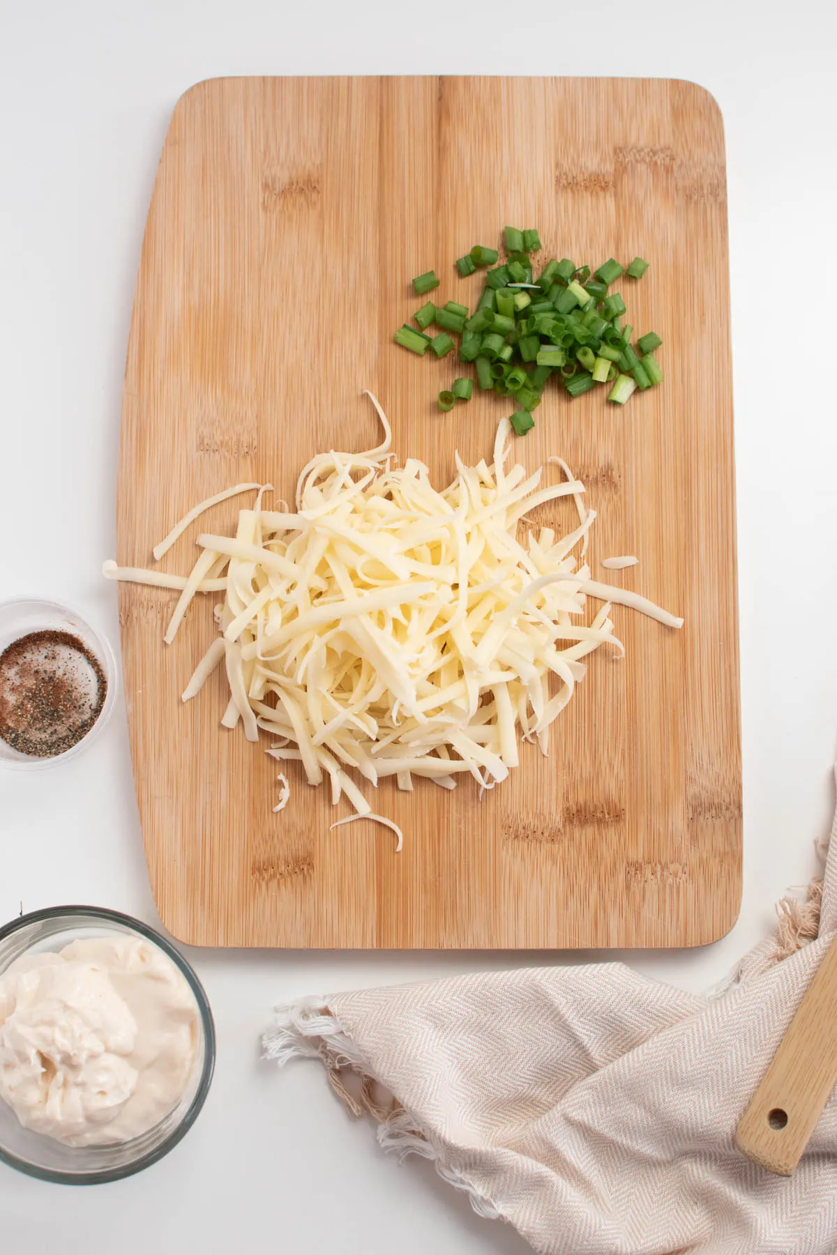 Piles of shredded Swiss cheese and chopped green onions on wood cutting board surrounded by bowls of ingredients.