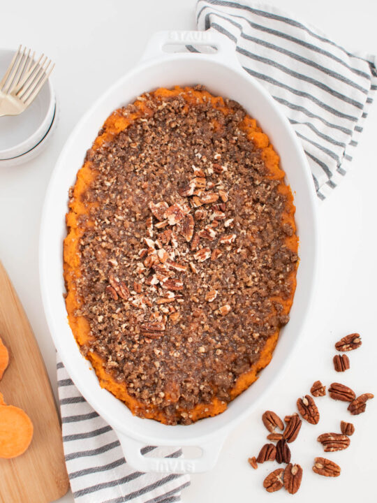 Sweet potato casserole with brown sugar crunch topping in white casserole dish surrounded by towel, pecans, and bowls.