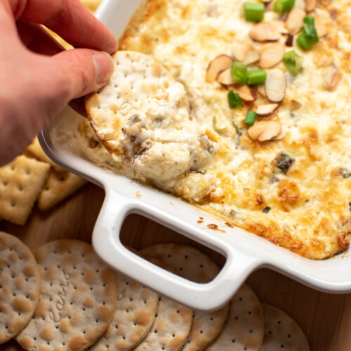 Hand dipping a cracker in a small baking dish of swiss almond dip.