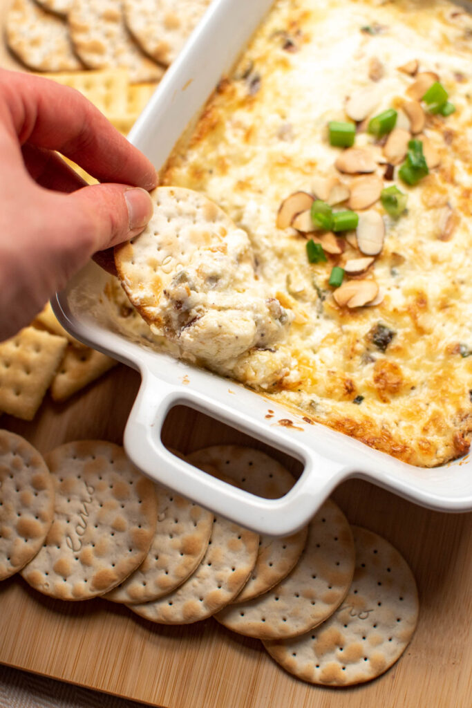 Hand dipping a cracker in a small baking dish of swiss almond dip.