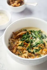 Creamed spinach with caramelized onions in white dinner bowl next to prep bowls with ingredients.