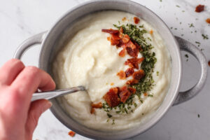 Woman stirs pot of creamy bacon cauliflower soup with green garnish and sprinkles of bacon.