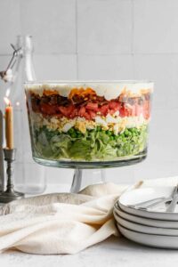 Glass trifle bowl with seven layer salad next to stack of plates.