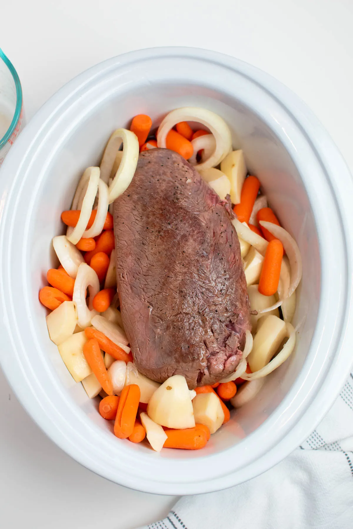 Browned roast in Crock Pot with raw onion, carrots and potato chunks all on white table.