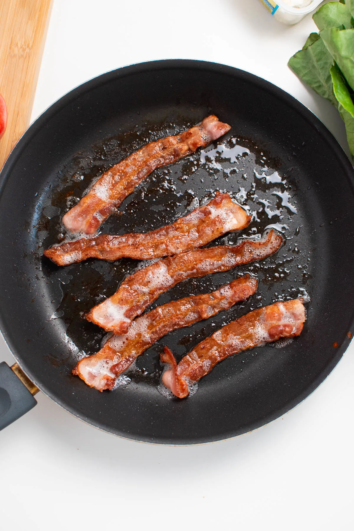 Five pieces of cooked bacon in black frying pan on white table.