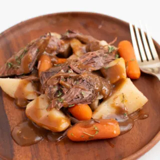 Close up of Crock Pot roast with carrots, potatoes, and gravy on wood plate.