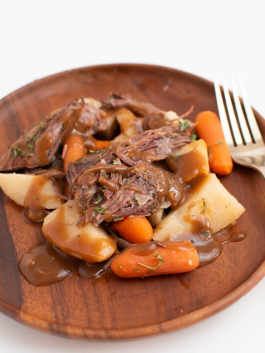 Close up of Crock Pot roast with carrots, potatoes, and gravy on wood plate.