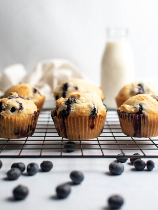 Fluffy blueberry muffins on baking rack with fresh blueberries scattered on counter.