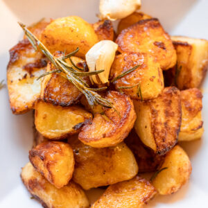 Duck fat roasted potatoes with cloves of garlic and green garnish in white bowl.
